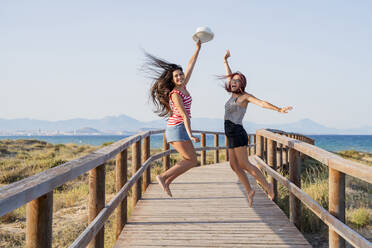 Excited teenage girls jumping on boardwalk at beach against clear sky - DLTSF00863