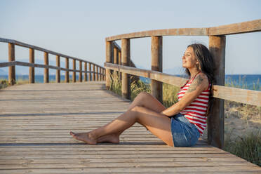 Thoughtful teenage girl sitting on boardwalk at beach against clear blue sky - DLTSF00865