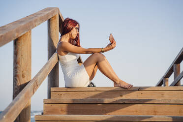 Teenager-Mädchen nimmt Selfie durch Smartphone, während auf hölzernen Stufen gegen klaren Himmel sitzen - DLTSF00872