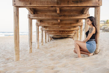 Thoughtful teenage girl sitting under pier at beach - DLTSF00874