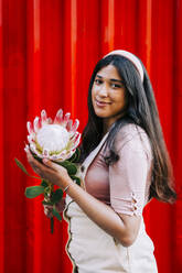 Junge Frau vor roter Wand, hält Protea-Blüte - DCRF00402