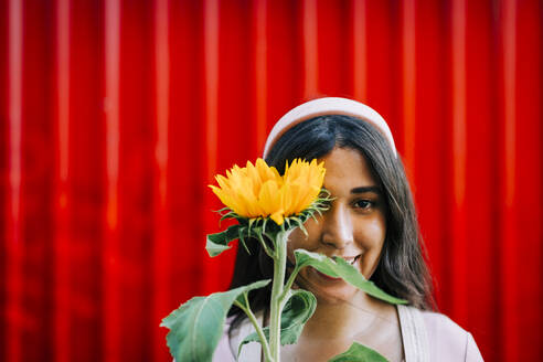 Junge Frau vor roter Wand, die ihr Auge mit einer Sonnenblume bedeckt - DCRF00408