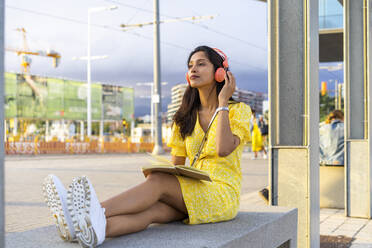 Young woman listening music while sitting on concrete bench in city - AFVF06775