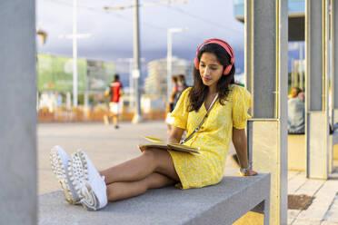 Young woman listening music while reading book on concrete bench in city - AFVF06776