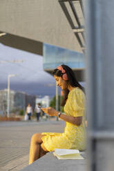 Young woman using smart phone and listening music while sitting on bench in city - AFVF06782