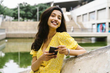 Happy woman holding smart phone while leaning on retaining wall in city - AFVF06794