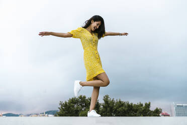 Carefree young woman standing with arms outstretched on retaining wall against sky - AFVF06801