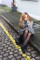 Nachdenkliche Frau in warmer Kleidung hält ihr Smartphone, während sie auf dem Bürgersteig sitzt - JMPF00159