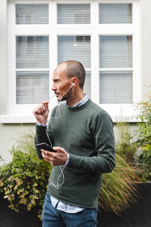 Mature man talking through in-ear headphones against window in city - JMPF00165