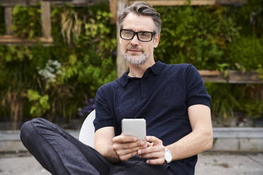 Nachdenklicher reifer Mann, der sein Smartphone hält, während er im Garten sitzt - SUF00635