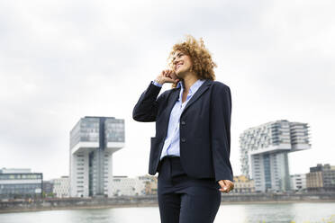 Smiling businesswoman talking over smart phone while standing against sky in city - MJFKF00488