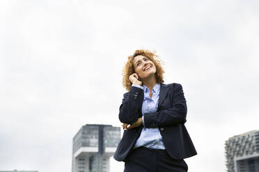 Smiling female entrepreneur talking over smart phone while standing against sky in city - MJFKF00489