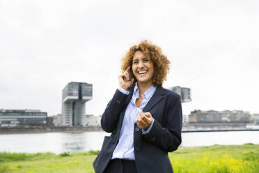 Fröhliche Unternehmerin mit lockigem Haar, die über ein Mobiltelefon spricht, während sie gegen den Himmel in der Stadt steht - MJFKF00490