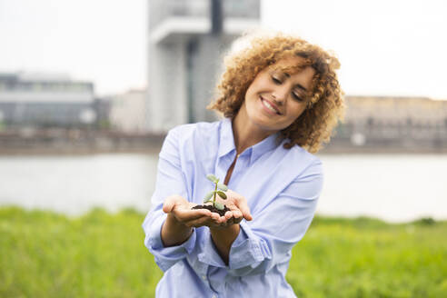 Lächelnde Geschäftsfrau mit lockigem Haar, die eine kleine Pflanze hält, während sie gegen den Himmel in der Stadt steht - MJFKF00498