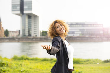 Smiling businesswoman with arm outstretched standing against river in city - MJFKF00506
