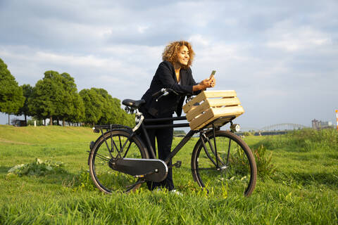 Businesswoman using smart phone while standing with bicycle on grassy land against sky stock photo