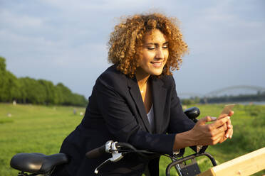 Lächelnde Geschäftsfrau mit lockigem Haar, die ein Smartphone benutzt, während sie mit dem Fahrrad vor dem Himmel steht - MJFKF00513