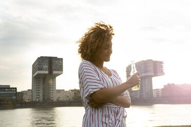 Mittlere erwachsene Frau, die eine Bierflasche hält, während sie vor einem Fluss und Gebäuden in der Stadt steht - MJFKF00515
