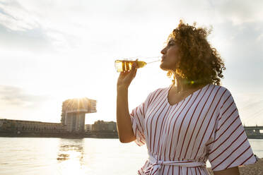 Mid erwachsene Frau mit lockigem Haar trinken Bier, während gegen den Himmel in der Stadt bei Sonnenuntergang stehen - MJFKF00521