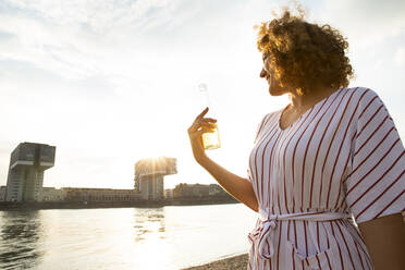 Lächelnde Frau mit lockigem Haar, die eine Bierflasche hält, während sie bei Sonnenuntergang auf einen Fluss in der Stadt blickt - MJFKF00522