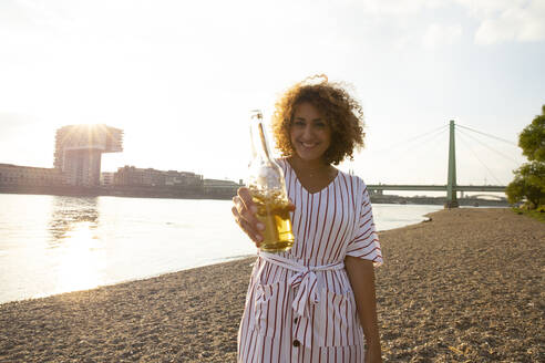 Glückliche Frau mit lockigem Haar, die eine Bierflasche in der Hand hält, während sie auf dem Land am Fluss in der Stadt steht - MJFKF00523