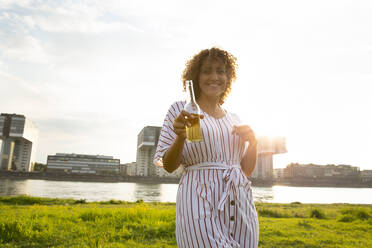 Happy woman with curly hair holding beer bottle while standing against river in city - MJFKF00524