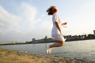 Mittlere erwachsene Frau mit Maske springt am Flussufer gegen den Himmel in der Stadt - MJFKF00526