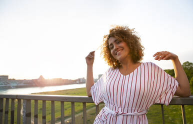 Smiling mid adult woman with curly hair leaning on railing against sky at sunset - MJFKF00527