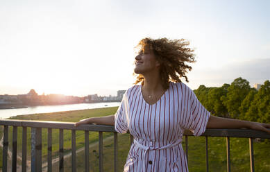 Lächelnde mittelgroße erwachsene Frau mit lockigem Haar, die an einem Geländer gegen den Himmel bei Sonnenuntergang steht - MJFKF00528