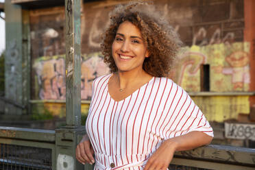 Cheerful woman with curly hair standing by railing against built structure during sunset - MJFKF00530