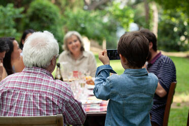 Boy photographing family at outdoor meal - CUF55780
