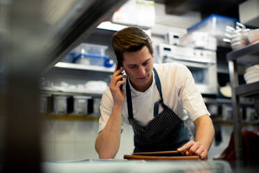 Junger männlicher Koch, der in der Küche ein digitales Tablet liest und mit seinem Smartphone spricht - CUF55801