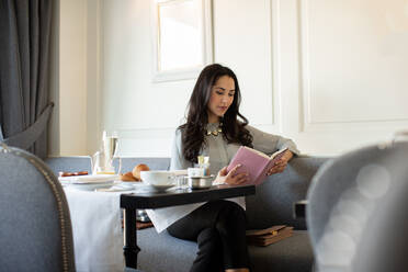 Junge Frau liest ein Buch und frühstückt mit Champagner in einem Boutique-Hotel in Italien - CUF55805