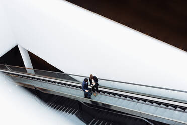 Geschäftsmann und Geschäftsfrau mit Gepäck auf Rädern auf einer Rolltreppe in einem Hotelgebäude - CUF55840