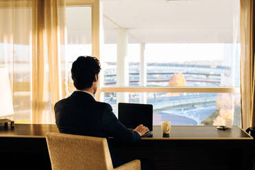 Businessman using laptop and looking out of hotel bedroom window - CUF55903