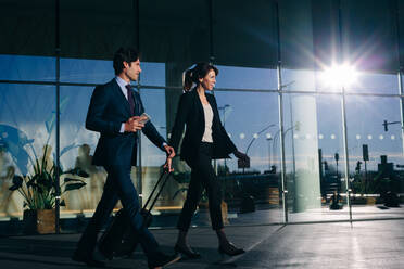 Businessman and businesswoman with wheeled luggage passing glass building, Malpensa, Milan - CUF55904