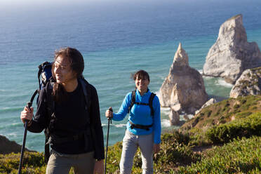 Two women with rucksacks and hiking poles walking along the coastline overlooking sea stacks. - CUF55913
