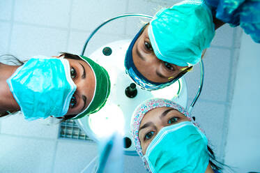 Blick von unten auf drei Chirurginnen mit OP-Masken, die in die Kamera schauen. - CUF55924
