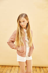 Porträt eines Mädchens mit langen blonden Haaren, das Shorts, Hemd und rosa Jacke trägt, auf blassgelbem Hintergrund. - CUF55937