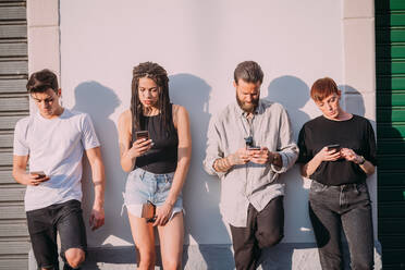 Two young women and men wearing casual clothes leaning against wall, using mobile phones. - CUF55943