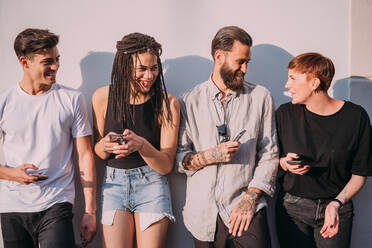 Two young women and men wearing casual clothes leaning against wall, using mobile phones. - CUF55944