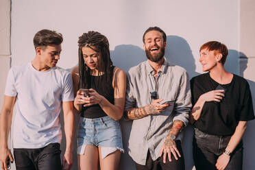 Two young women and men wearing casual clothes leaning against wall, using mobile phones. - CUF55945