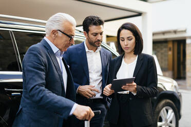 Zwei Geschäftsleute und eine Frau vor einem Hotel mit Blick auf ein digitales Tablet - CUF55975