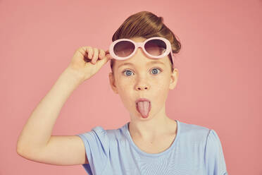 Porträt eines brünetten Mädchens mit blauem T-Shirt und Sonnenbrille auf rosa Hintergrund, das der Kamera die Zunge entgegenstreckt. - CUF56003