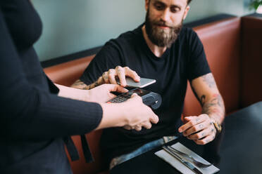 High angle close up of tattooed bearded man sitting in a bar, using mobile phone to pay. - CUF56015