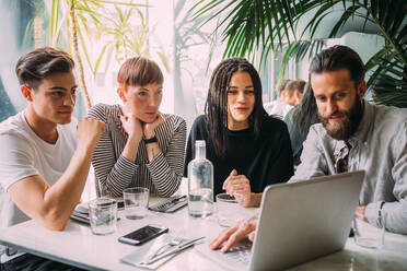 Zwei junge Frauen und Männer in Freizeitkleidung sitzen an einem Tisch in einer Bar und schauen auf einen Laptop. - CUF56032