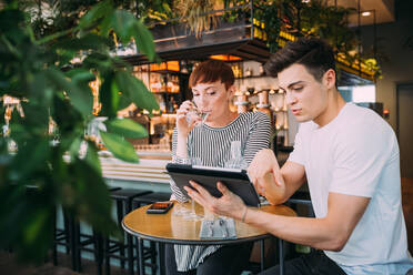 Junge Frau und Mann sitzen an einem Tisch in einer Bar und schauen auf ein digitales Tablet. - CUF56036