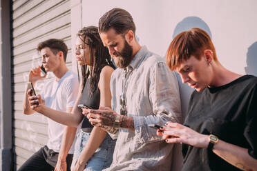 Two young women and men wearing casual clothes leaning against wall, using mobile phones. - CUF56048