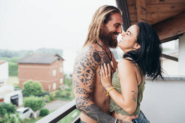 Bärtiger, tätowierter Mann mit langen brünetten Haaren und Frau mit langen braunen Haaren stehen auf einem Balkon, umarmen und küssen sich. - CUF56054