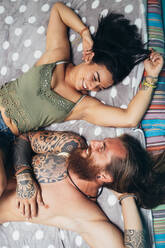 Ein bärtiger, tätowierter Mann mit langen brünetten Haaren und eine Frau mit langen braunen Haaren liegen auf einem Bett und lächeln sich gegenseitig an. - CUF56056